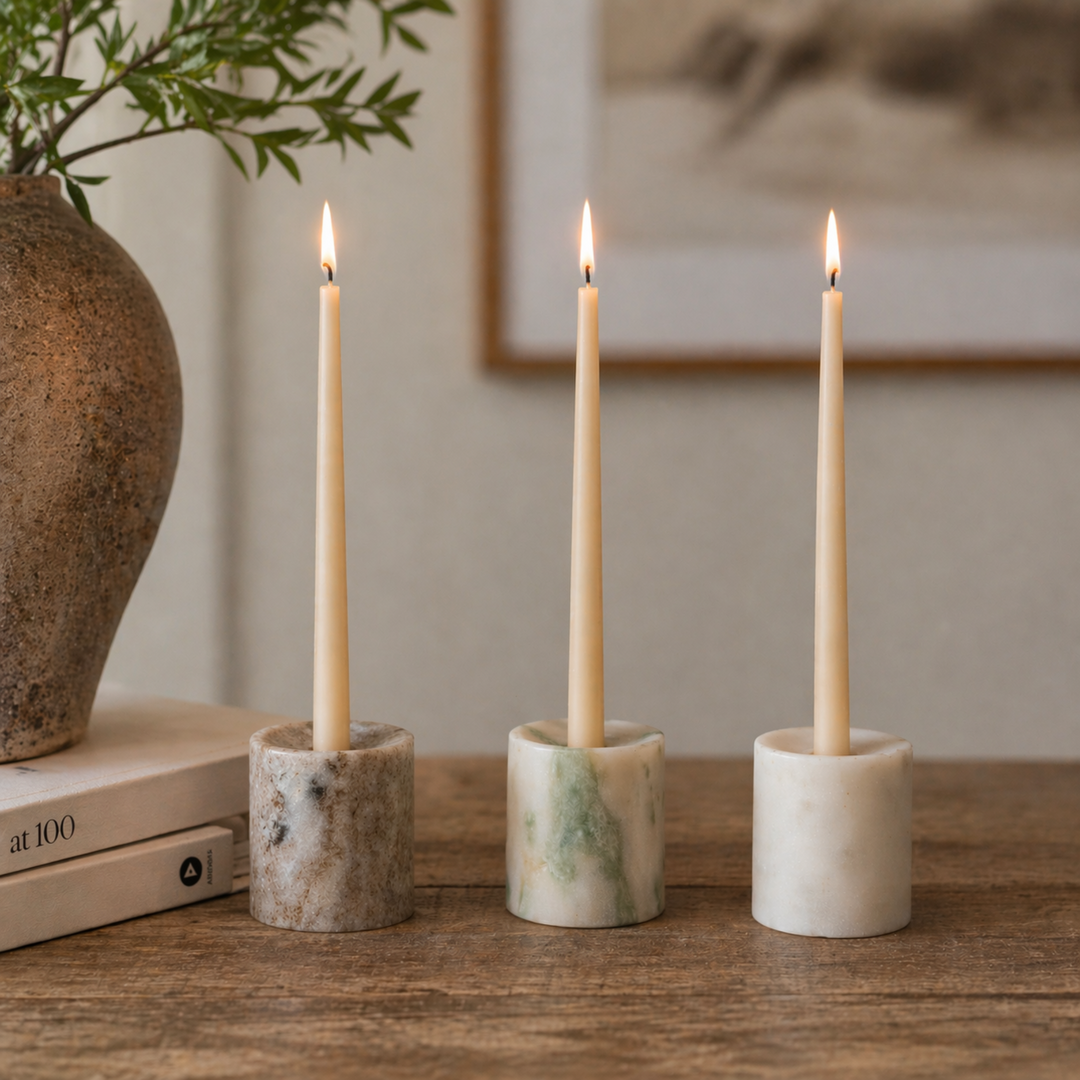 Three lit candles in marble holders on a wooden surface with a vase and book in the background.