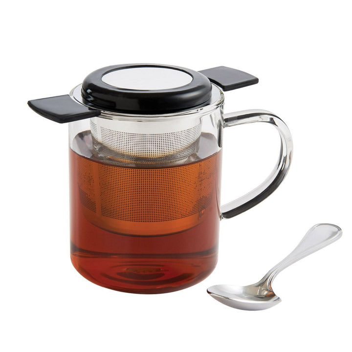 Glass teapot with black lid and handle, filled with tea, next to a silver spoon on a white background.