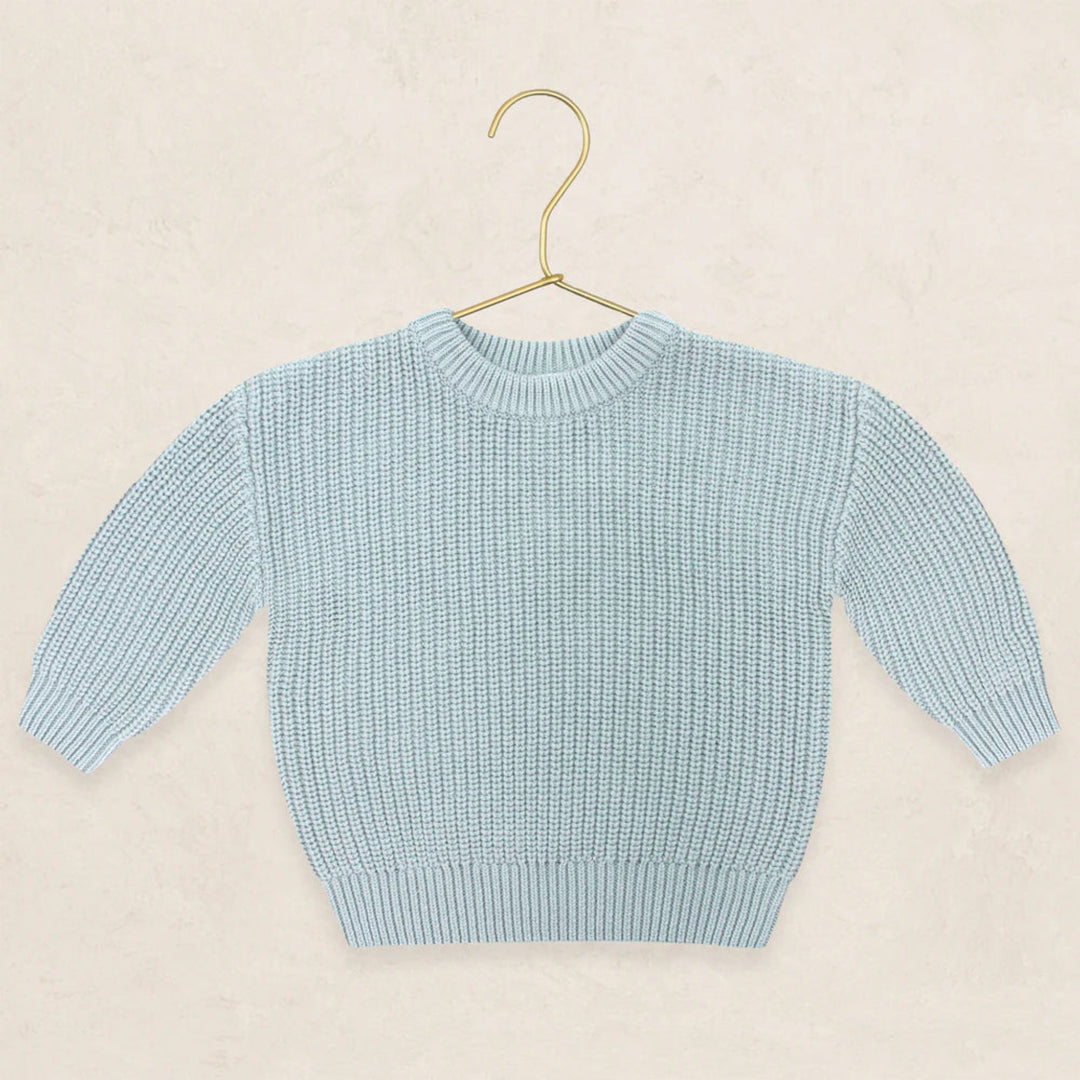 A light blue ribbed knit sweater with long sleeves and a crew neck, displayed on a gold metal hanger against a textured beige background.