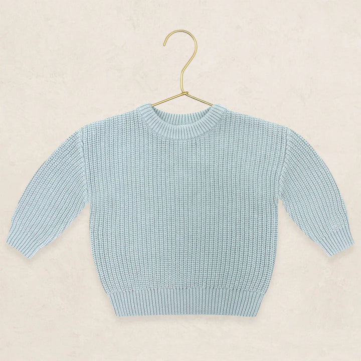 A light blue ribbed knit sweater with long sleeves and a crew neck, displayed on a gold metal hanger against a textured beige background.