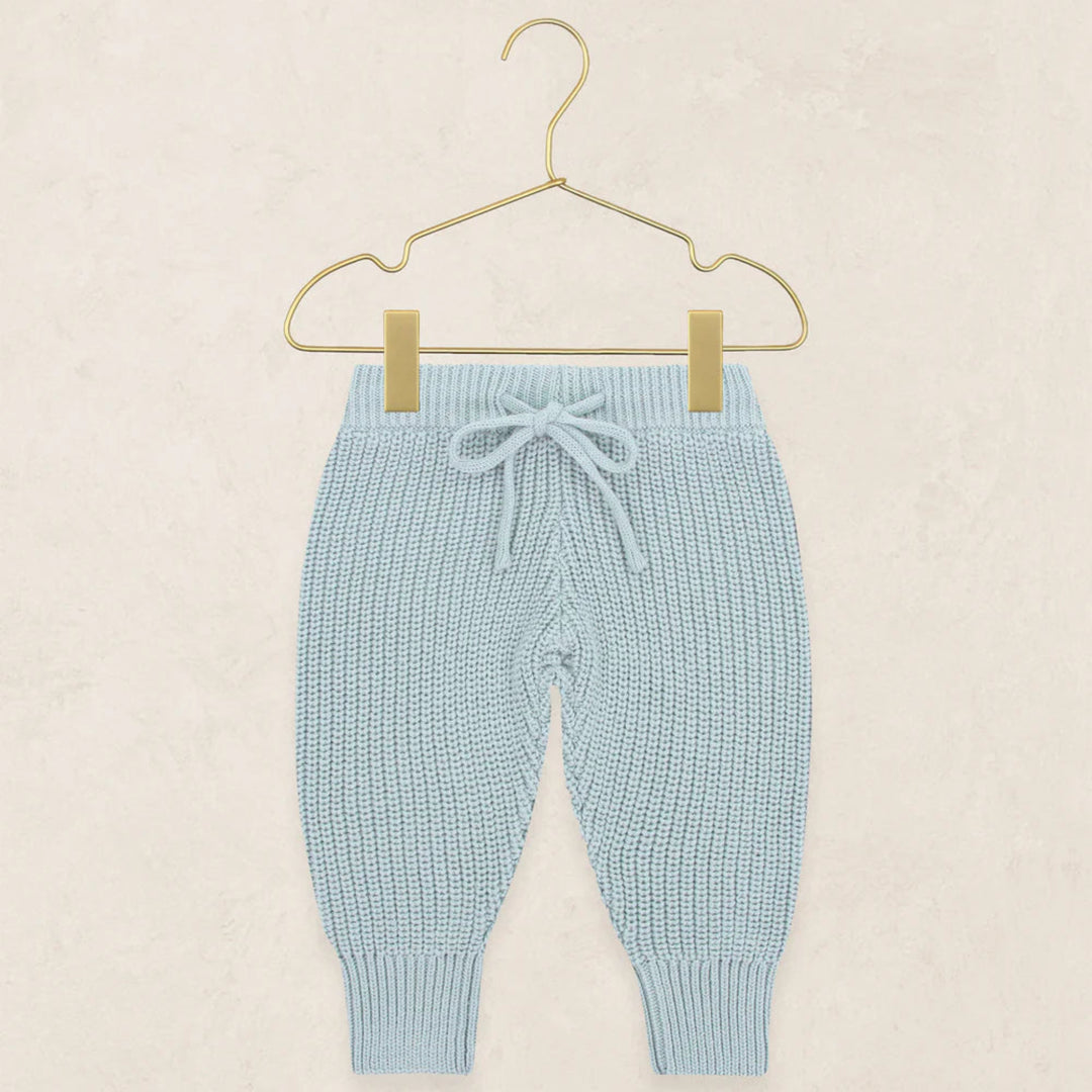 Light blue knit baby pants with a drawstring waist and ribbed ankle cuffs, hanging from a gold wire hanger against a textured beige background.