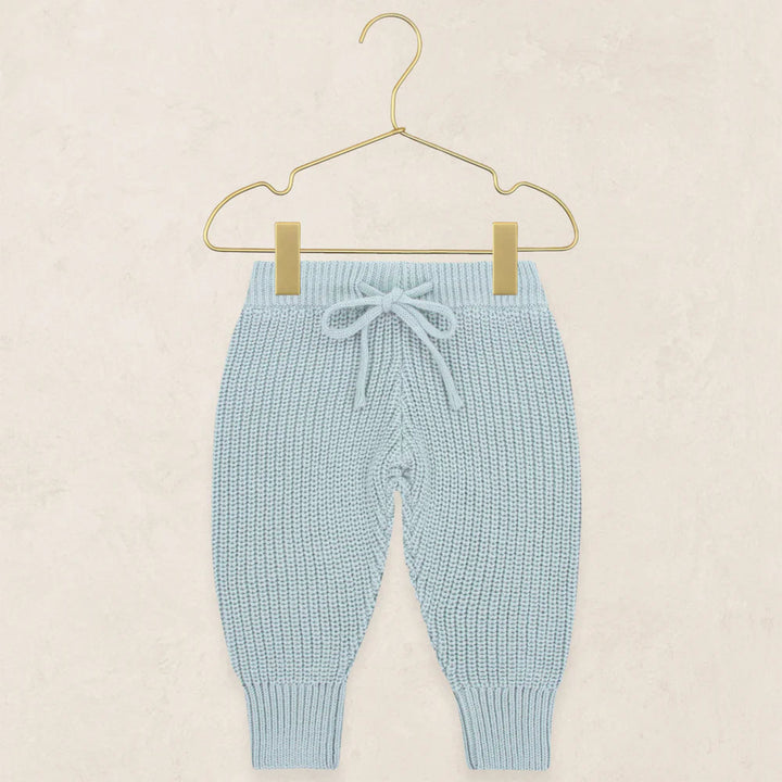 Light blue knit baby pants with a drawstring waist and ribbed ankle cuffs, hanging from a gold wire hanger against a textured beige background.