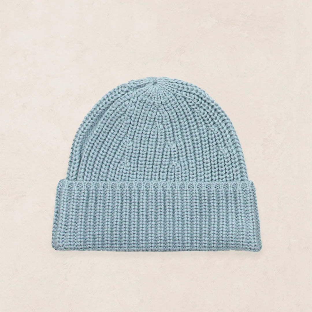 A light blue knit beanie hat with a ribbed cuff, displayed flat against a light beige textured background.