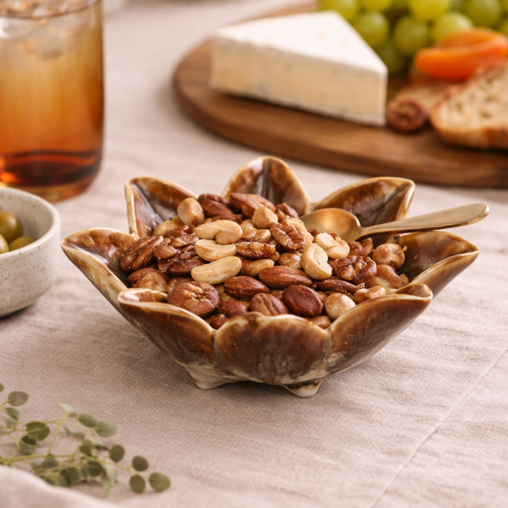 Nuts in a decorative bowl on a table with a glass of iced tea and cheese platter in the background.