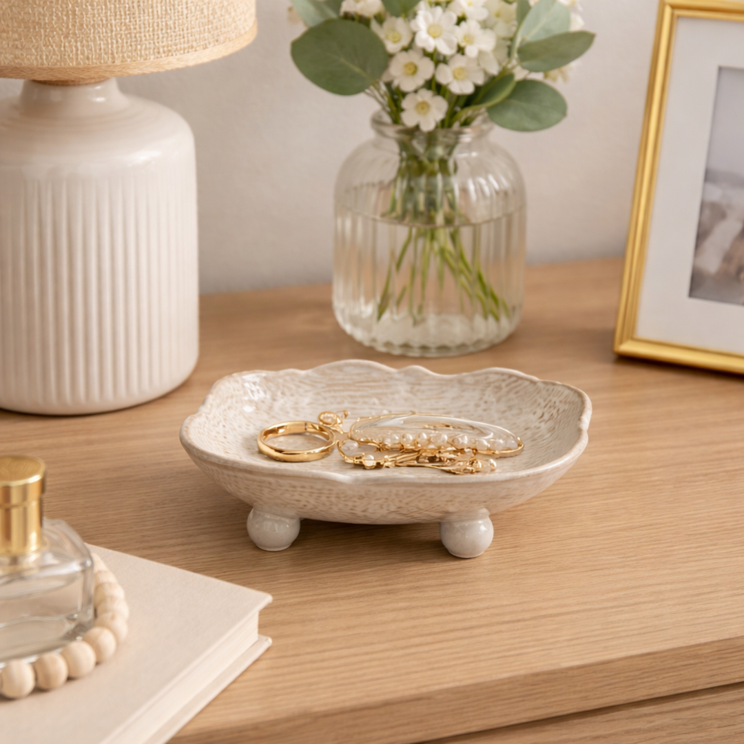 Decorative jewelry dish with gold rings on a wooden surface with a lamp, flowers, and photo frame in the background.
