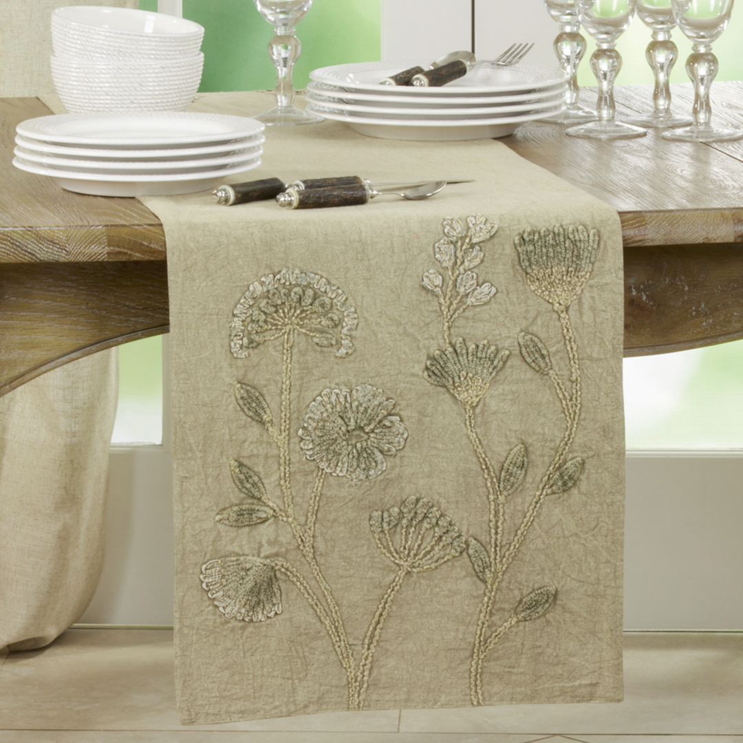 Dining table with a floral embroidered table runner and white plates and glasses.