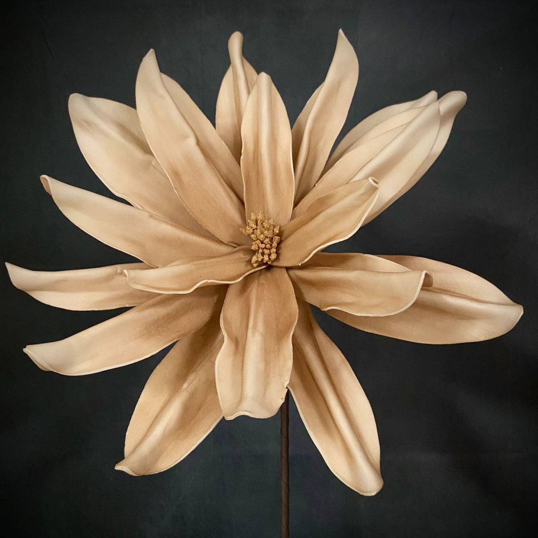 A single large artificial flower with pale beige petals and a small tan center, mounted on a dark stem against a dark charcoal background.
