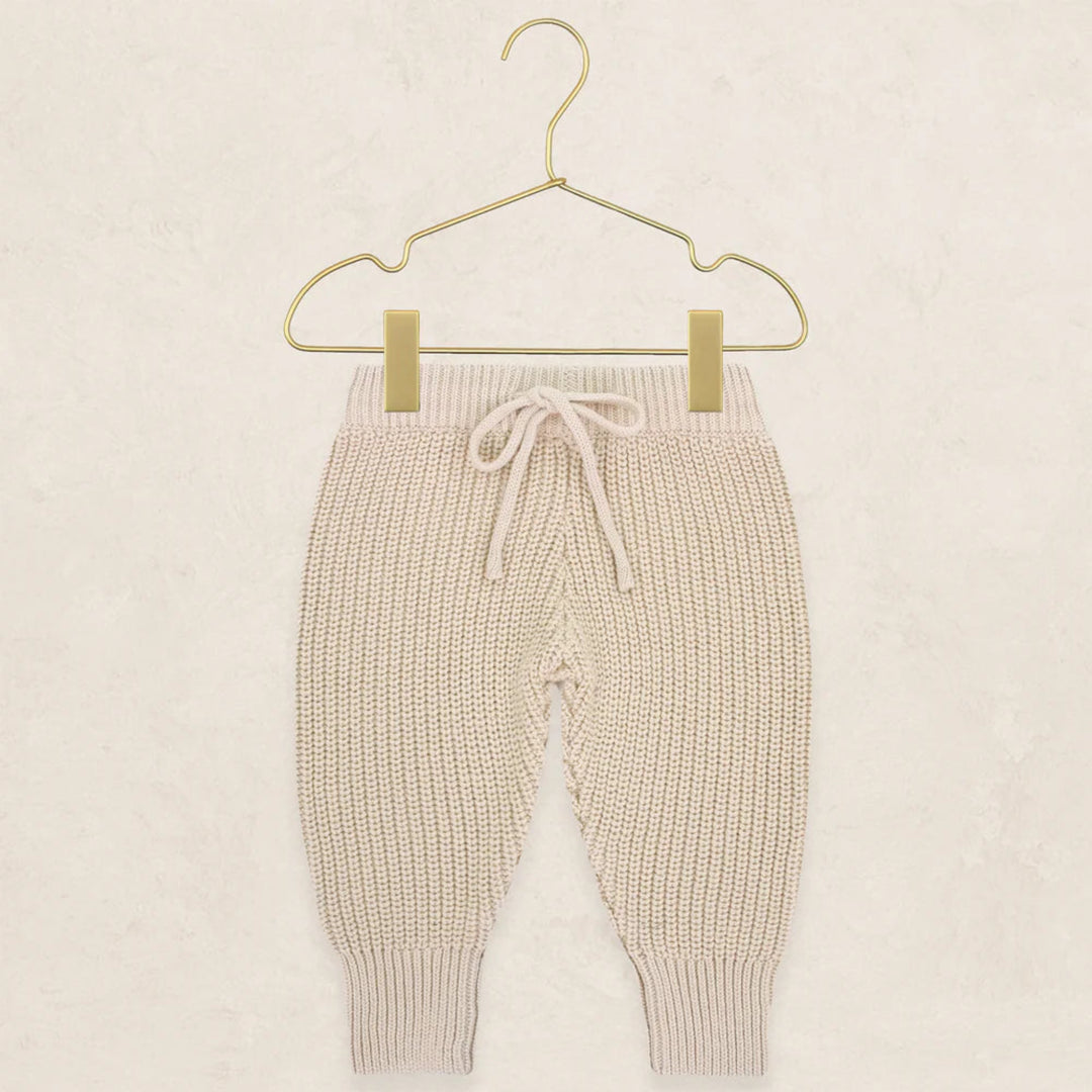 Cream-colored ribbed knit baby pants with a drawstring waist, hanging from a gold metal hanger against a textured beige background.