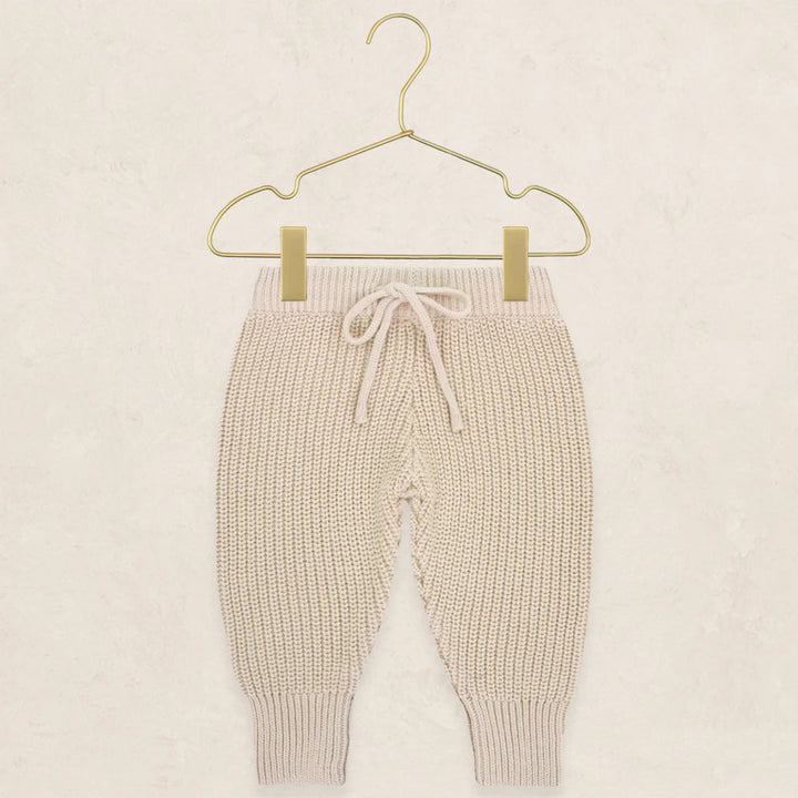 Cream-colored ribbed knit baby pants with a drawstring waist, hanging from a gold metal hanger against a textured beige background.
