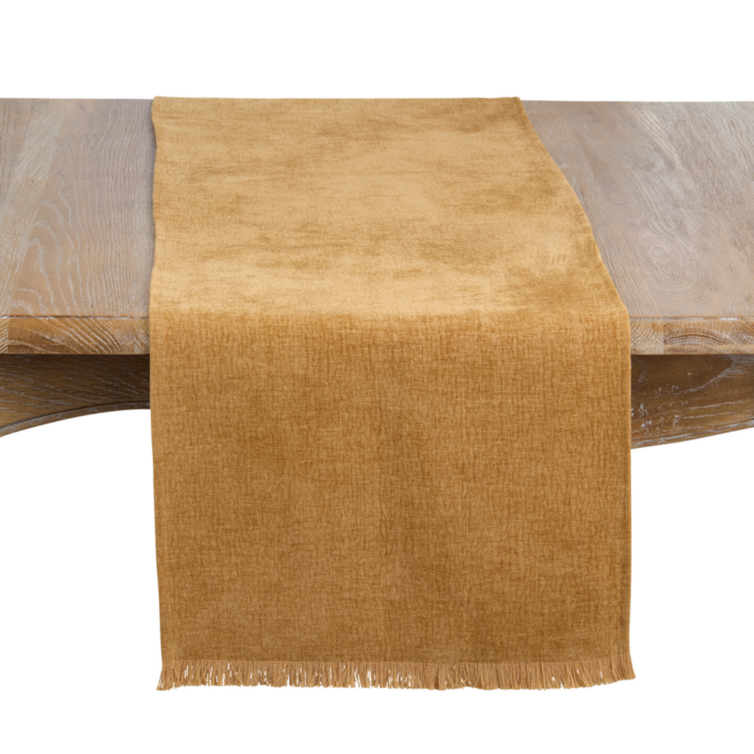 A tan woven table runner with fringe, draped over a wooden table against a white background.