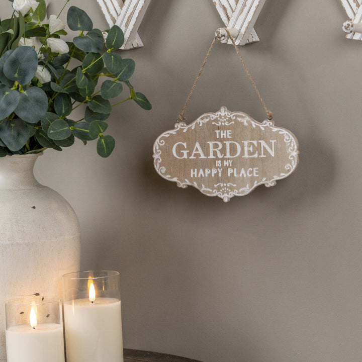 Decorative sign with 'The Garden is My Happy Place' on a wall with candles and a plant.