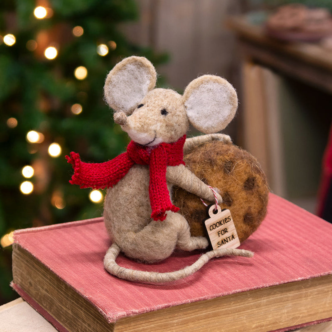 Felt mouse with a red scarf and cookie, sitting on a book with a Christmas tree in the background.