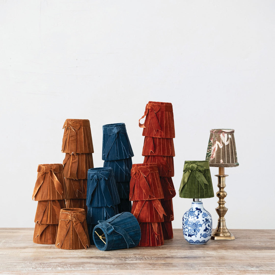 A group of decorative table lamps with tiered fabric shades in brown, blue, red, green, and metallic gold, displayed on a light wood surface against a plain white wall.
