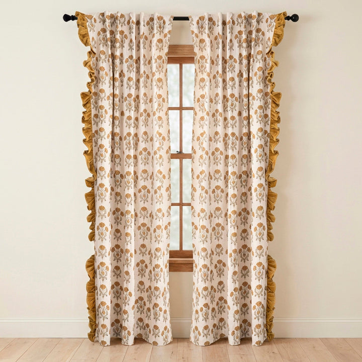 A pair of floor-length curtain panels with a cream background, a repeating tan damask-style floral pattern, and a wide ruffled trim along the edges, hanging on a black rod over a window.