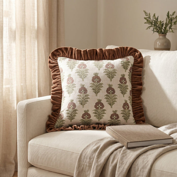 A decorative throw pillow with a brown ruffled border and a cream floral pattern, styled on a beige sofa with a throw blanket and a book.