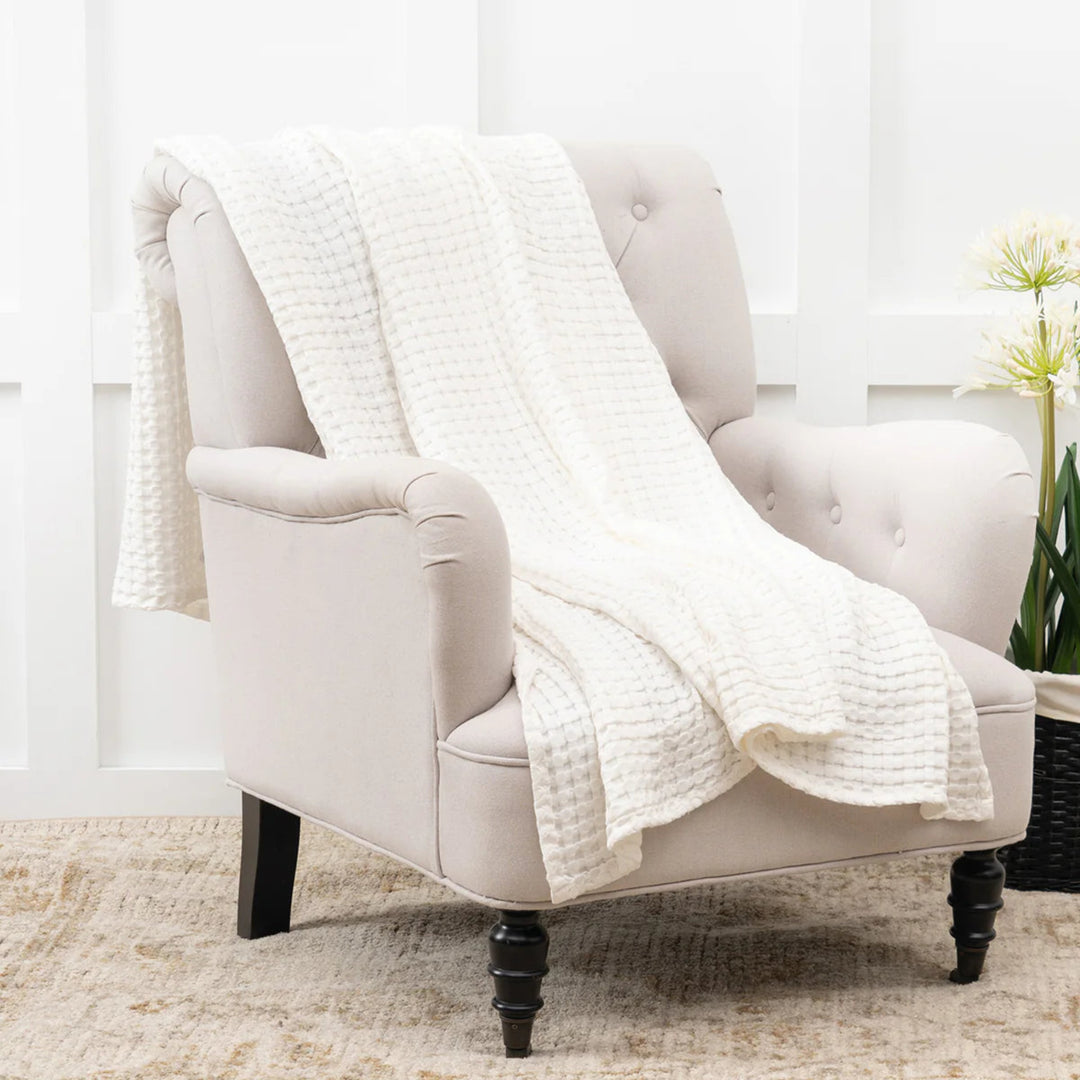 White armchair with a textured white blanket draped over it, set against a neutral background.