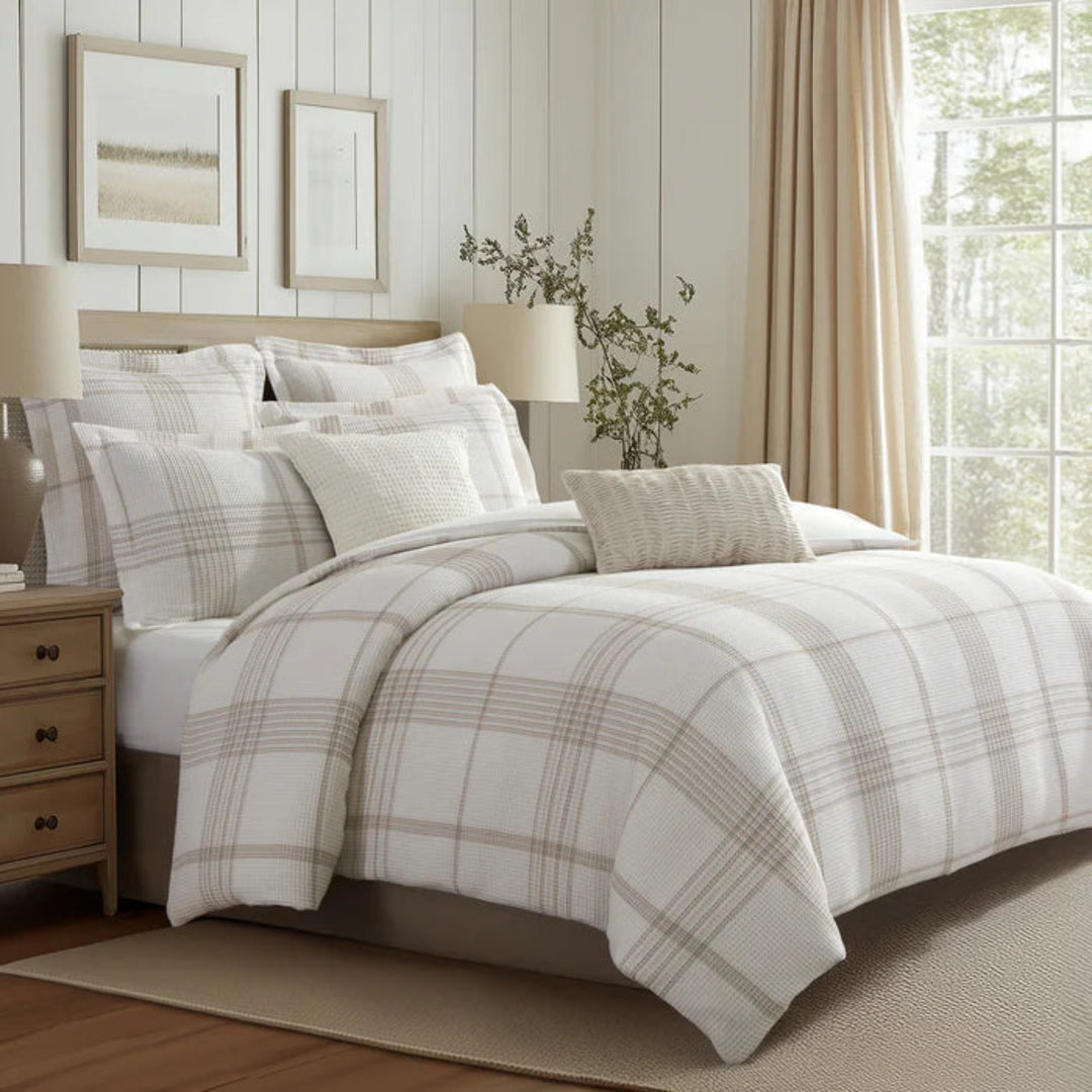 A bed dressed in a cream and beige plaid comforter set with matching shams and accent pillows, styled in a bright bedroom with wood paneling, a window with beige curtains, and bedside tables with lamps.