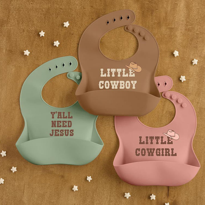 Three silicone baby bibs in sage green, brown, and dusty pink are arranged on a textured tan background with small white star confetti. The bibs feature western-themed text and graphics, including 'LITTLE COWBOY', 'Y'ALL NEED JESUS', and 'LITTLE COWBOY GIRL' with cowboy hat illustrations.