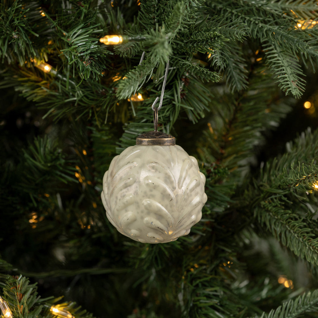 A textured, off-white pinecone-shaped Christmas ornament hanging from a green artificial Christmas tree with warm yellow lights.