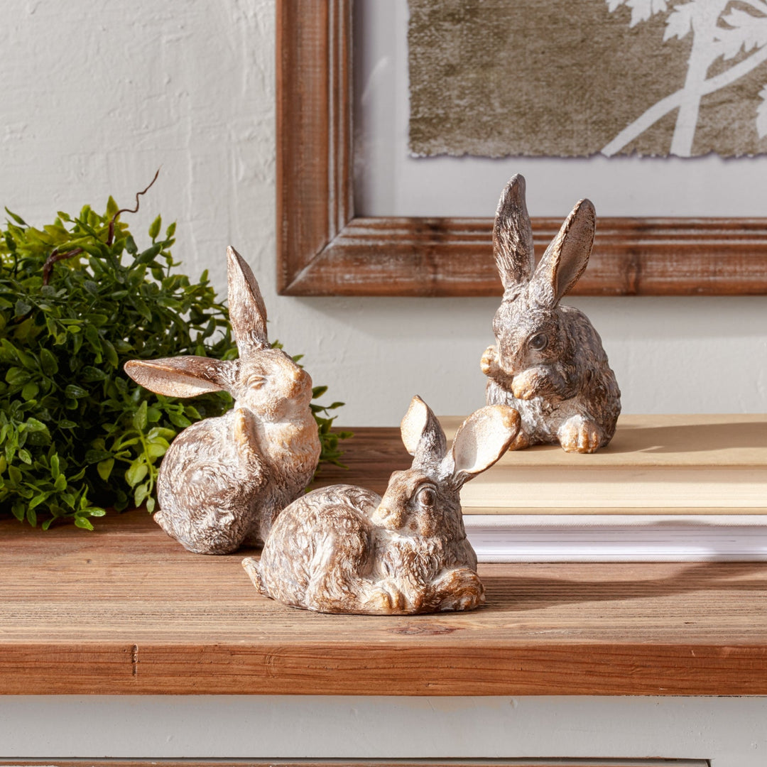 Three decorative rabbit figurines on a wooden surface with a framed picture in the background.