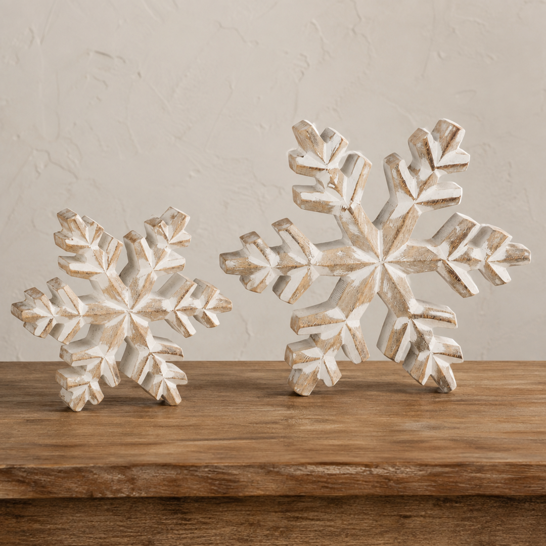 Two distressed white wooden snowflake ornaments displayed on a rustic wooden surface against a textured beige wall.