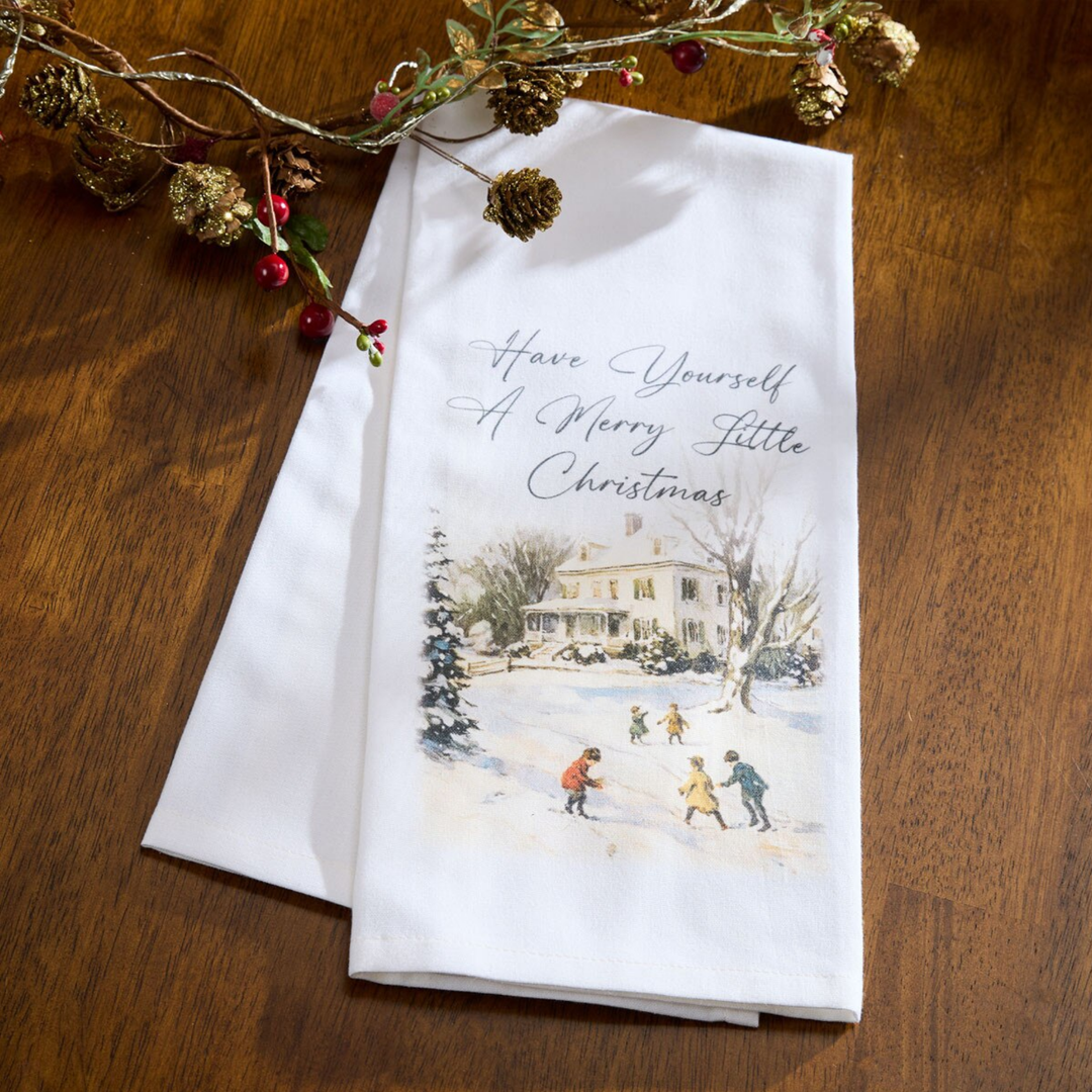 A white kitchen towel with a festive illustration of children ice skating in front of a house, printed with the text 'Have Yourself A Merry Little Christmas'. The towel is displayed on a dark wooden table alongside a holiday pine cone garland.