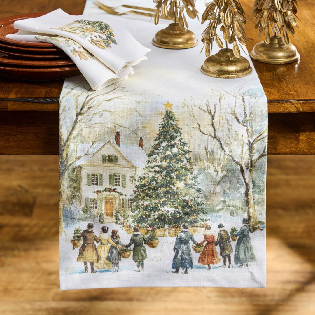 Decorative table runner with a winter scene of a house and people gathered around a Christmas tree on a wooden table.