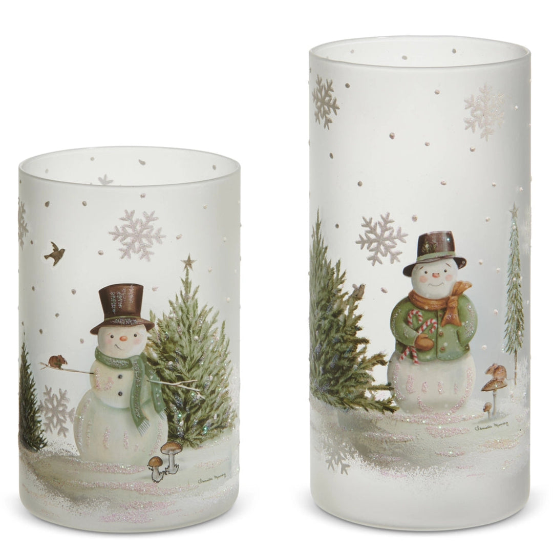 Two cylindrical glass candle holders with winter-themed designs featuring snowmen and trees on a white background.