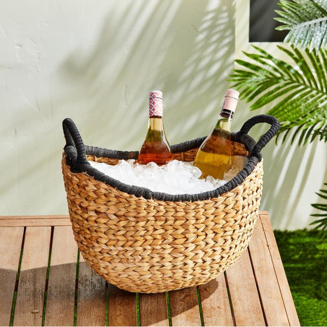 Wicker ice bucket with bottles of wine on a wooden table outdoors.