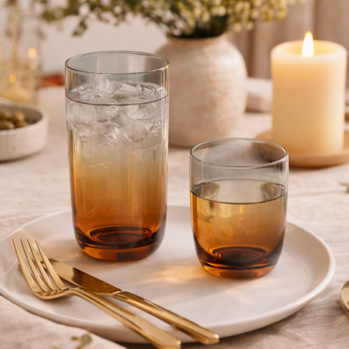 Two amber/green iridescent drinking glasses on a white plate with gold cutlery, set on a table with candles and flowers.