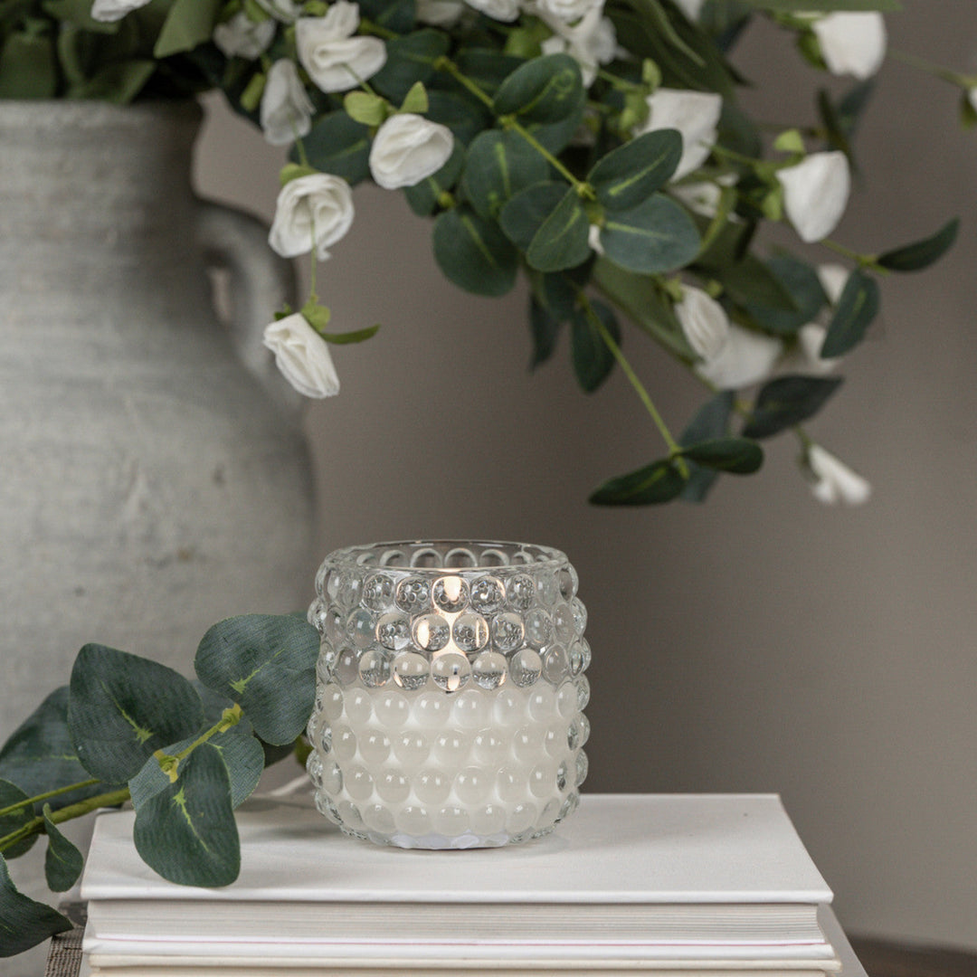 Clear textured candle holder on a white surface with greenery and a vase in the background