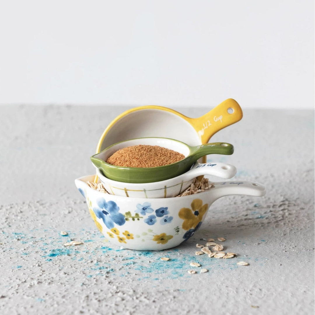 Three stacked ceramic measuring cups with a floral pattern, shown on a textured light gray surface with oats and blue powder scattered around them.