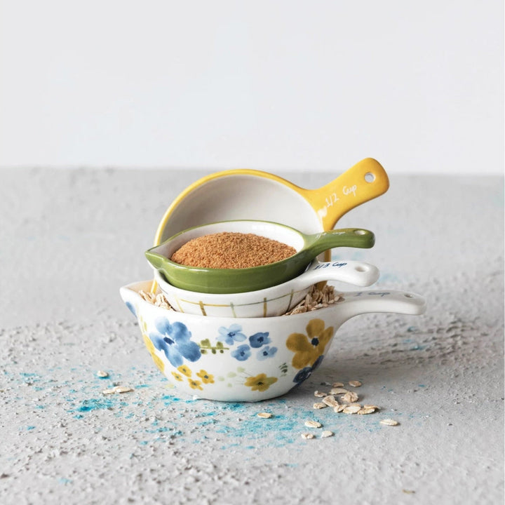 Three stacked ceramic measuring cups with a floral pattern, shown on a textured light gray surface with oats and blue powder scattered around them.